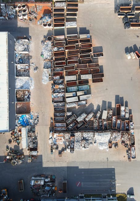 Aerial view of an outdoor storage yard showing rows of rusted metal bathtubs, sinks, and other discarded household fixtures arranged in columns on a concrete surface. To the left, there are pallets with various packaging materials and pile of broken items, with some objects covered in blue tarps. A large grey building borders the yard on the left side. In the lower part of the image, a small vehicle and a forklift are present, indicating equipment used for loading and transportation. The site is illuminated by natural daylight, highlighting the metallic textures and shadows cast by objects. This outdoor depot appears to be part of a waste or scrap collection facility or a recycling site, relevant to large-scale household item disposal or home relocation services conducted by Man with Van Malden Rushett, as shown in the context of furniture transport and packing during removals.