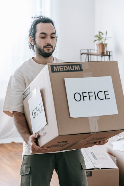 A white moving van with the words 'Moving Company Local & Long Distance' printed on its side is parked on a street in front of a brick building with large, rectangular windows. The van's driver, weari