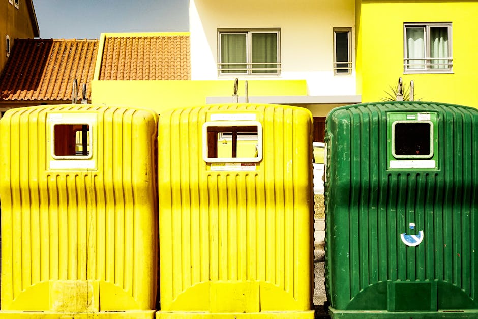 Three large waste disposal containers are positioned outdoors in front of a residential building. Two yellow recycling bins are placed side by side, with small rectangular openings at the top for waste insertion and vented ribbed plastic exteriors. To the right, a green container with a similar vented design and a small circular recycling sticker is visible. The background shows a modern house with white walls and multiple windows, one of which has a small balcony. The area appears to be part of a home relocation or waste collection process, with the containers likely used for disposing of household waste or recyclable materials. The scene is well-lit, highlighting the vibrant colors of the bins and the building facade, with a focus on the logistics of waste collection in the context of moving or house clearance, as handled by Man with Van Malden Rushett.