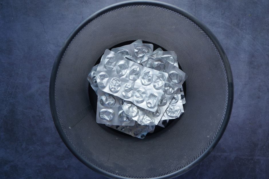An overhead view of a round, black metal trash bin filled with crumpled aluminum foil food packaging and a plastic tray, positioned on a concrete floor near a doorway. In the background, part of a wooden doorframe and a grey skirting board are visible, suggesting the bin is located inside a home or property during a house removal or packing process. Smaller pieces of packaging are also visible beneath the foil, indicating recent packing or disposal activity typically associated with household moving and packing procedures. This scene reflects the routine of clearing waste materials in the context of home relocation services provided by Man with Van Malden Rushett, supporting efficient furniture transport and packing efforts.