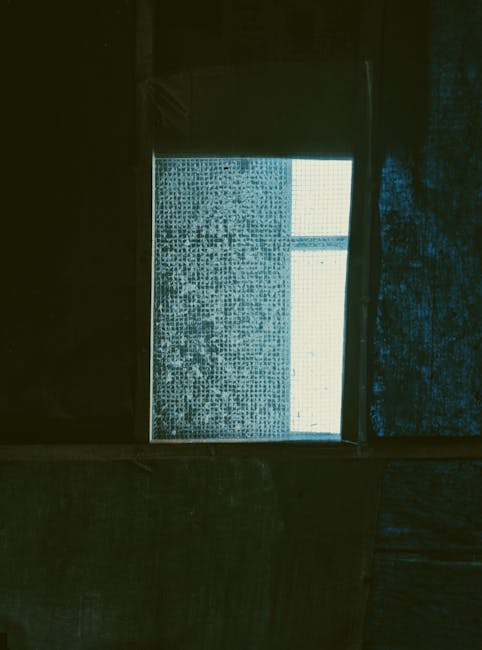 A photograph taken from inside a building showing a window with a frosted or patterned glass pane, partially covered by a dark curtain or blind on the right side. The window frame is visible, with a clear rectangular section in the top right corner allowing light to enter the room. The environment appears to be a residential interior, possibly a living room or hallway, with dim lighting and textured wall surfaces surrounding the window. The scene is neutral and does not depict any furniture, boxes, or moving equipment, but it provides context for the natural environment during a house removal or furniture transport process, as discussed on the page about fragile-item insurance for Malden Rushett moves.
