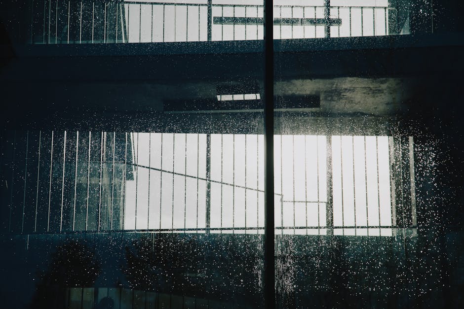 The image shows a large glass window with rain droplets on its surface during daylight. Behind the glass, there is a view of an indoor space with visible metal railings and a ceiling with industrial-style lighting fixtures. The window reflects the interior environment, highlighting the presence of structural elements typical in buildings preparing for a house removal or moving process. The lighting suggests natural daylight illuminating the scene, with the overall setting indicating an environment of a property being prepared for a home relocation or furniture transport, consistent with professional removals services by Man with Van Malden Rushett. This visual aligns with the context of packing and moving logistics, illustrating the physical environment involved in furniture transport and the careful handling required during a property move.
