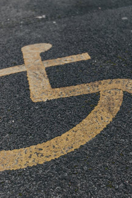Close-up view of a yellow disabled parking space symbol painted on an asphalt surface outside a residential property. The symbol, consisting of a stylized chair with a backrest and a person sitting, appears worn with some fading and minor cracks. The surrounding area shows rough-textured dark asphalt with small pebbles and gravel embedded in the surface. In the background, part of a driveway or pavement is visible, leading to the entrance of a property where furniture, cardboard boxes, and packing materials may be staged for a home relocation or furniture transport. This scene reflects the preparation phase of a house move, where vehicles such as a van from Man with Van Malden Rushett are likely involved in loading furniture, appliances, and boxes, using equipment like trolleys, straps, or blankets. The natural outdoor lighting indicates daytime, with no people present in the image, focusing on the parking marking and surface condition relevant to house removals and movement logistics.