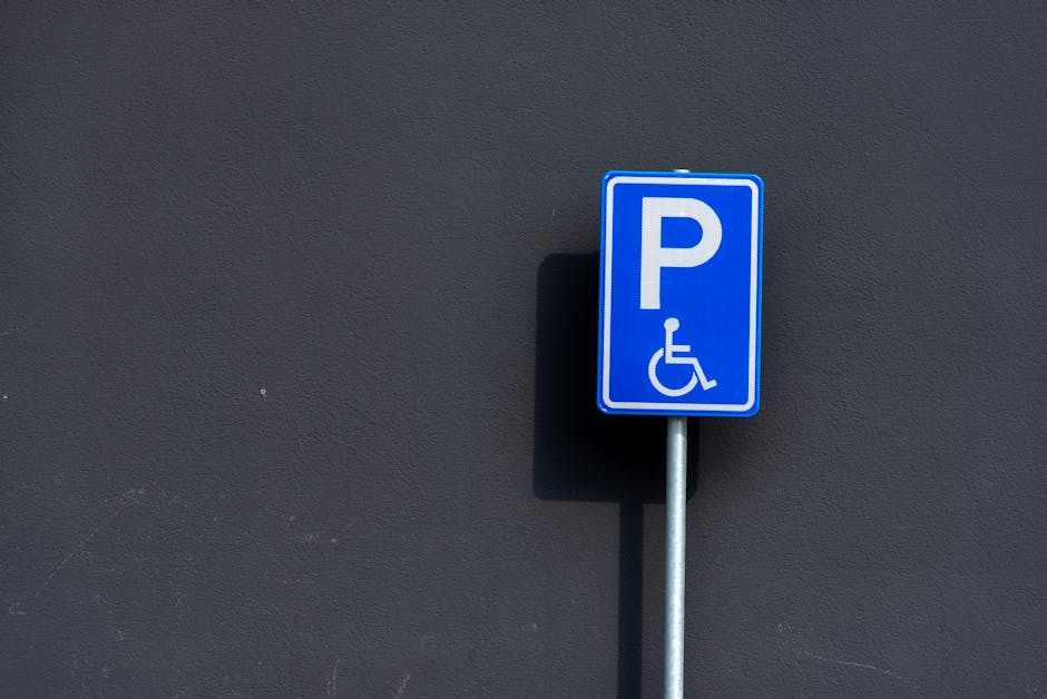 A blue rectangular parking sign with a white uppercase letter 'P' and a white wheelchair symbol below it, indicating accessible parking, mounted on a silver metal pole positioned against a dark grey exterior wall. The sign casts a subtle shadow onto the wall. The environment suggests an outdoor setting adjacent to a property entrance or driveway, with no additional objects visible. This image relates to house removals and moving logistics by illustrating accessible parking options for loading or unloading furniture and boxes during home relocation, as part of the services offered by Man with Van Malden Rushett.