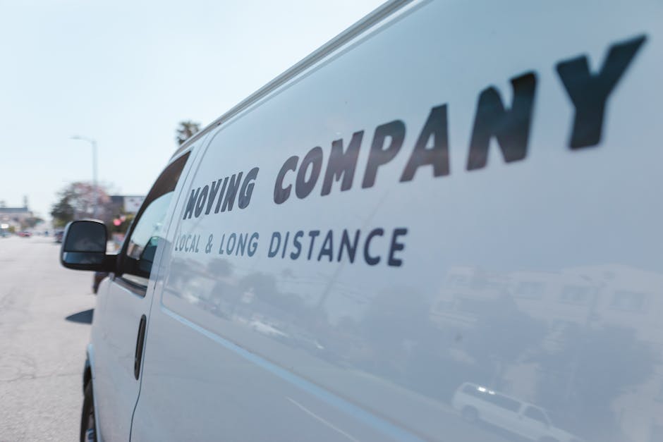 Close-up of a white moving van parked outdoors in a parking lot with a clear sky, displaying the partial logo 'MOVING COMPANY' along with the text 'LOCAL & LONG DISTANCE' in bold dark letters. The van is positioned near the curb, with the driver's side mirror visible on the left. In the background, there are other parked vehicles, buildings, and street signs, indicating a typical urban or suburban environment. The image captures the van during the loading process for a home relocation, with the focus on the vehicle used by Man with Van Malden Rushett for furniture transport and packing and moving services.