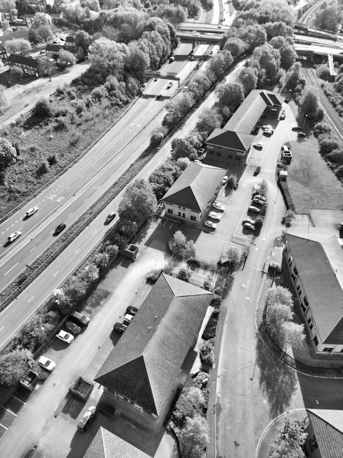 An aerial black-and-white photograph showing a residential area with multiple buildings and houses, paved pathways, and a parking lot filled with cars. The scene captures a loading process involving a man with a van, visible on the street adjacent to the houses, assisting with the transportation of furniture and boxes wrapped in plastic and blankets. The van is parked beside the property, with some items, including cardboard boxes and furniture wrapped in protective materials, being moved or prepared for loading. Surrounding the buildings are trees and greenery, with a dual carriageway visible in the background, along with roadside vegetation and highway overpasses. The natural lighting and shadows indicate daytime, highlighting the organized home relocation and furniture transport activities typical of a same-day moving service provided by Man with Van Malden Rushett, as part of their house removals and packing and moving operations.
