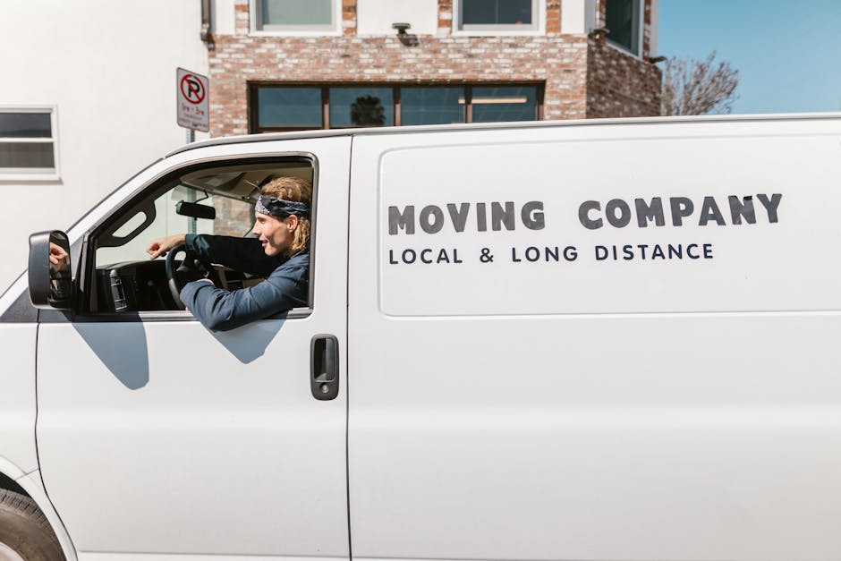 Close-up of a white moving van parked outdoors in a parking lot with a clear sky, displaying the partial logo 'MOVING COMPANY' along with the text 'LOCAL & LONG DISTANCE' in bold dark letters. The van is positioned near the curb, with the driver's side mirror visible on the left. In the background, there are other parked vehicles, buildings, and street signs, indicating a typical urban or suburban environment. The image captures the van during the loading process for a home relocation, with the focus on the vehicle used by Man with Van Malden Rushett for furniture transport and packing and moving services.
