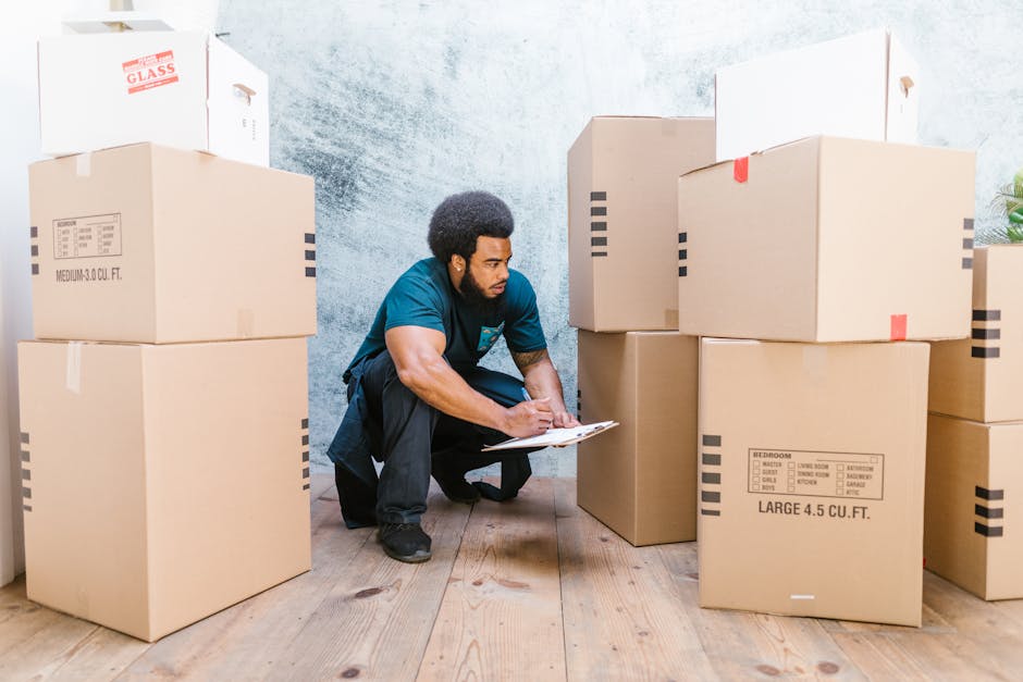 A man with dark, curly hair and a beard, wearing a teal T-shirt and black pants, is positioned on one knee inside a room, taking notes on a clipboard. Surrounding him are several large cardboard boxes labeled with descriptions of contents such as bedroom items, and measurements like 'LARGE 4.5 CU.FT.' These boxes are stacked on a wooden floor near a light grey wall. Some boxes have red or white tape sealing them, indicating packing for a home relocation. A white box with red text marked 'GLASS' is visible on top of a stack, suggesting fragile items. The scene depicts an interior space prepared for moving or furniture transport, with the man possibly coordinating or inspecting the packing process. This aligns with furniture transport, packing and moving activities, as part of professional removals services provided by Man with Van Malden Rushett.