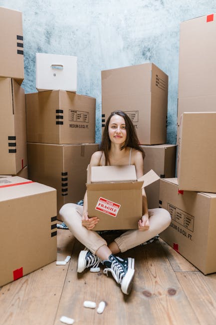 A young woman sits on a wooden floor inside a room surrounded by numerous cardboard boxes, some sealed with red and black packing tape, and a few labeled with red FRAGILE stickers. She holds an open box on her lap, and there are small packing materials, such as foam peanuts, scattered nearby. The background features a textured light blue wall, and the scene is well-lit with natural or artificial light, illustrating a home relocation or packing process alongside furniture and moving preparations. The setting reflects the packing stage of a house move, with the presence of various unpacked boxes and the woman's calm expression. This image aligns with furniture transport and moving services provided by Man with Van Malden Rushett.