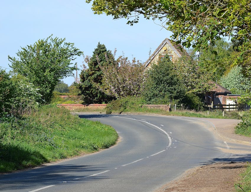 A quiet residential road in Malden Rushett with a gently curving asphalt surface marked by dashed white lines in the centre. On the left side of the road, there is a grassy verge with overgrown shrubs and small wildflowers. To the right, a narrow sidewalk runs alongside the verge, partially shaded by a large leafy tree extending branches overhead. In the background, there is a traditional, pitched-roof house partially obscured by dense greenery, including a tall conifer and several blossoming trees with pink and white flowers. The scene is illuminated by natural daylight under a clear blue sky, indicating a typical peaceful suburban area suitable for home relocation or furniture transport, often supported by local removals services such as Man with Van Malden Rushett. The overall environment suggests a quiet, rural village road optimized for safe loading and unloading of household items, with no vehicles or movement visible, emphasizing a calm setting for home packing and moving activities.